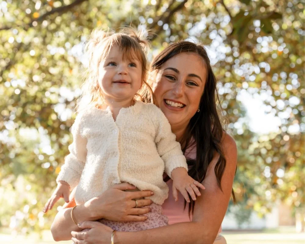 A woman holding a young girl outdoors on a sunny day, smiling and surrounded by trees with autumn leaves.