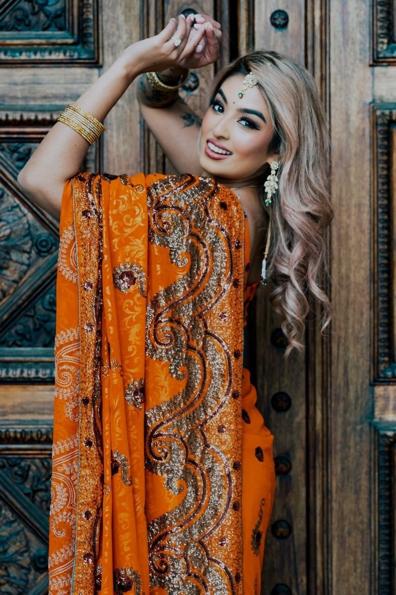 A woman in a vibrant orange traditional dress with intricate gold and silver embroidery, smiling and posing indoors in front of a decorative wooden door with ornate carvings.