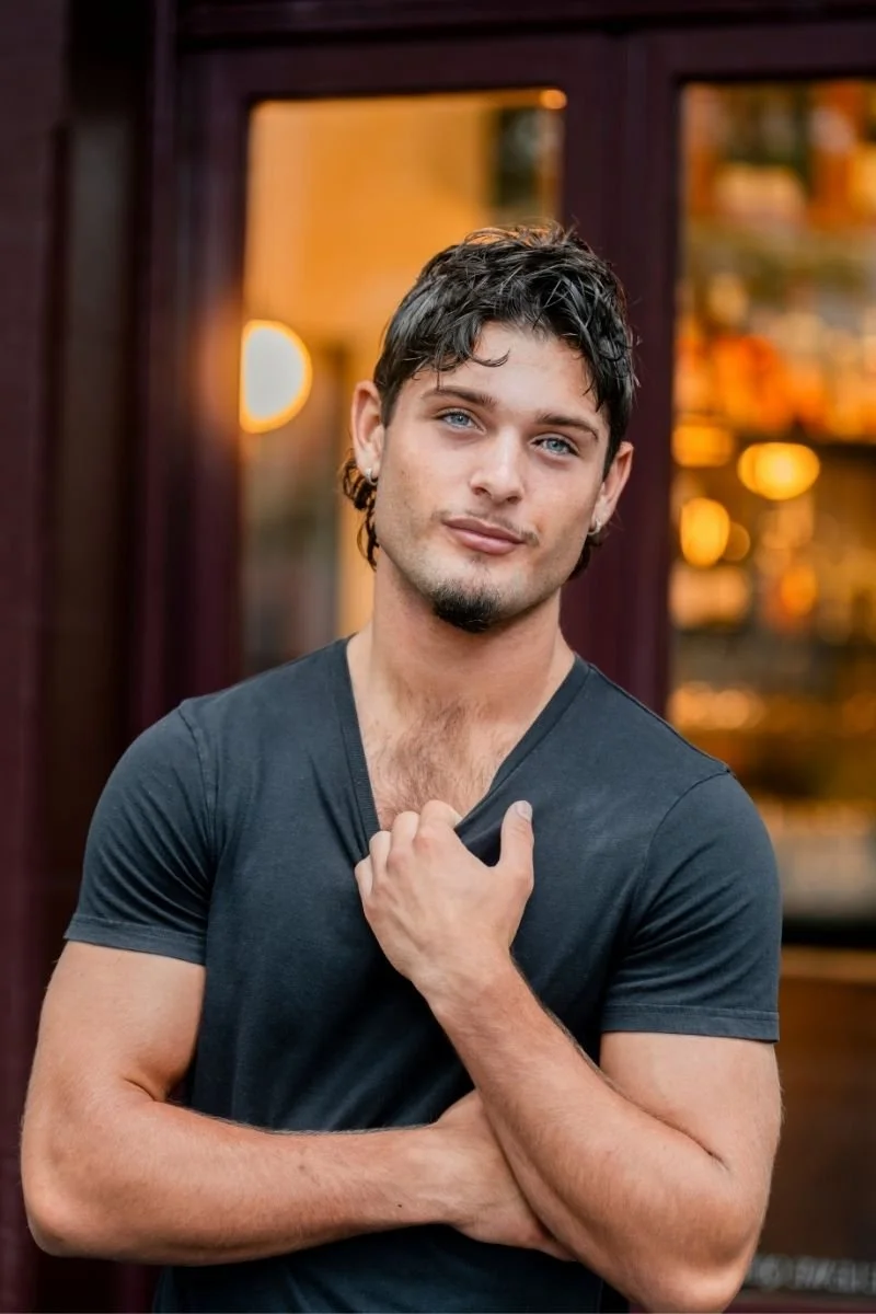 Young man with dark hair, light skin, and blue eyes standing outside in front of a window with warm lighting, wearing a dark V-neck T-shirt, with one hand on his chest and the other crossing his body.