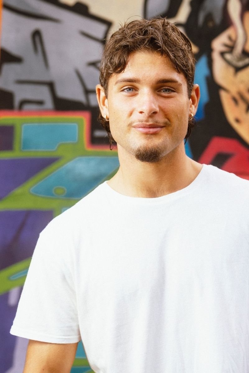 A young man with short, wavy brown hair, a light beard and mustache, and wearing a white t-shirt standing in front of a colorful graffiti wall.