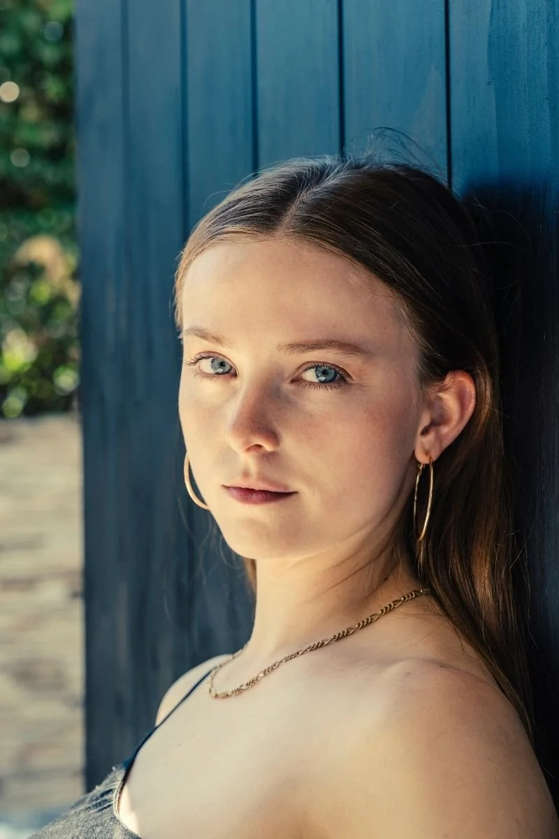Close-up of a young woman with long brown hair and blue eyes, wearing gold hoop earrings and a gold necklace, leaning against a dark blue wooden wall outdoors.