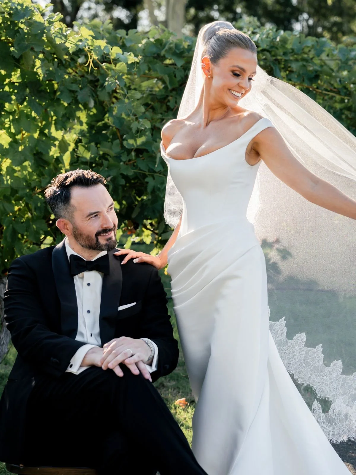 A bride in a white wedding gown with a veil stands next to a groom in a black tuxedo with a bow tie, posing outdoors with a backdrop of greenery.
