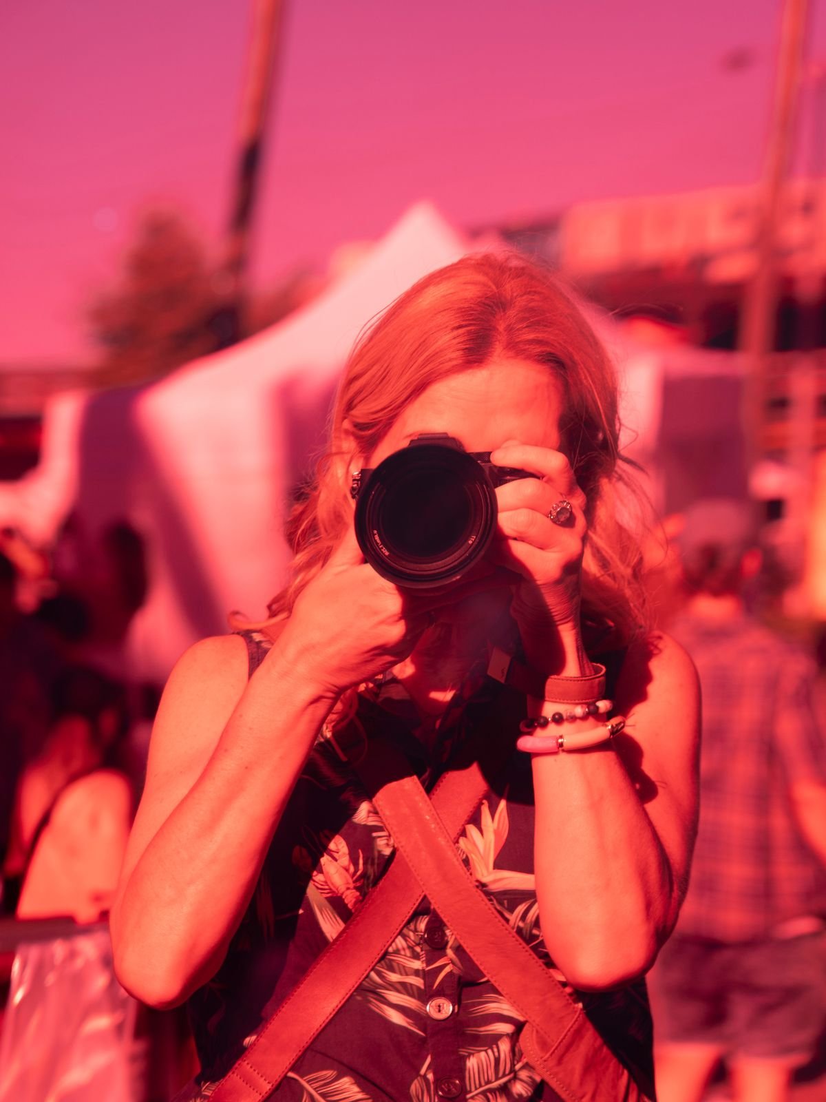 A woman with shoulder-length hair taking a photograph with a camera at an outdoor event during sunset, with a pinkish sky in the background.