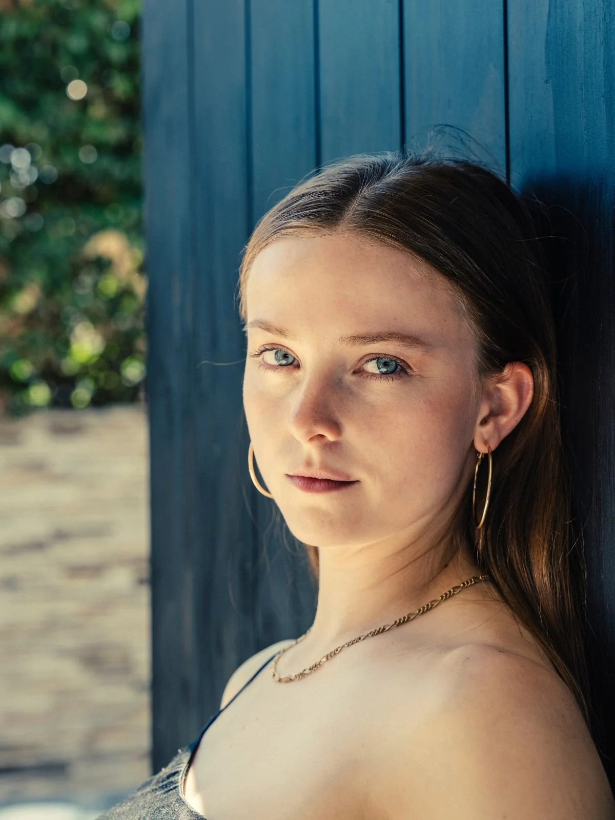A young woman with blue eyes and long brown hair, posing against a dark blue wooden wall outdoors.