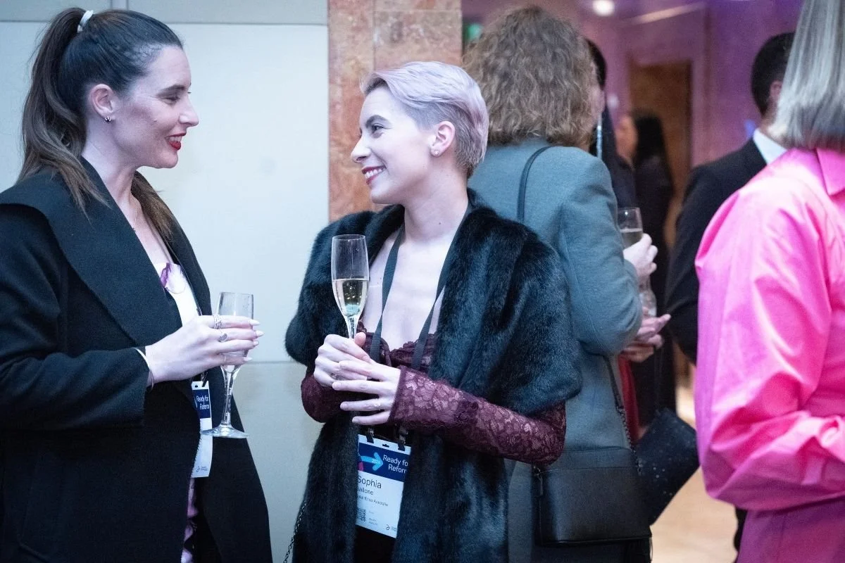 Two women conversing at a social event, holding glasses of champagne, all dressed in formal attire.