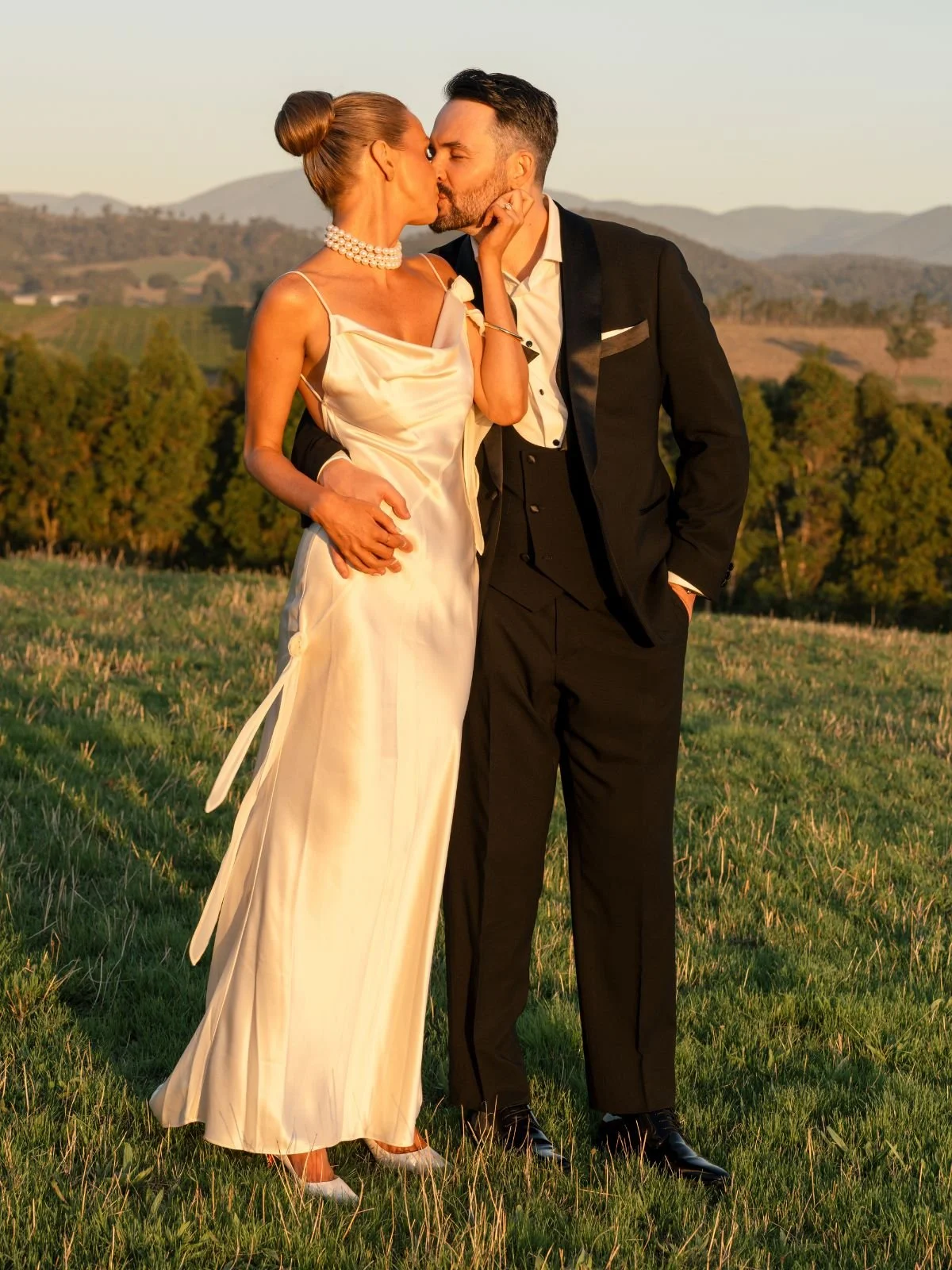 A couple dressed in formal wedding attire, standing outdoors on a grassy hill at sunset, sharing a kiss.