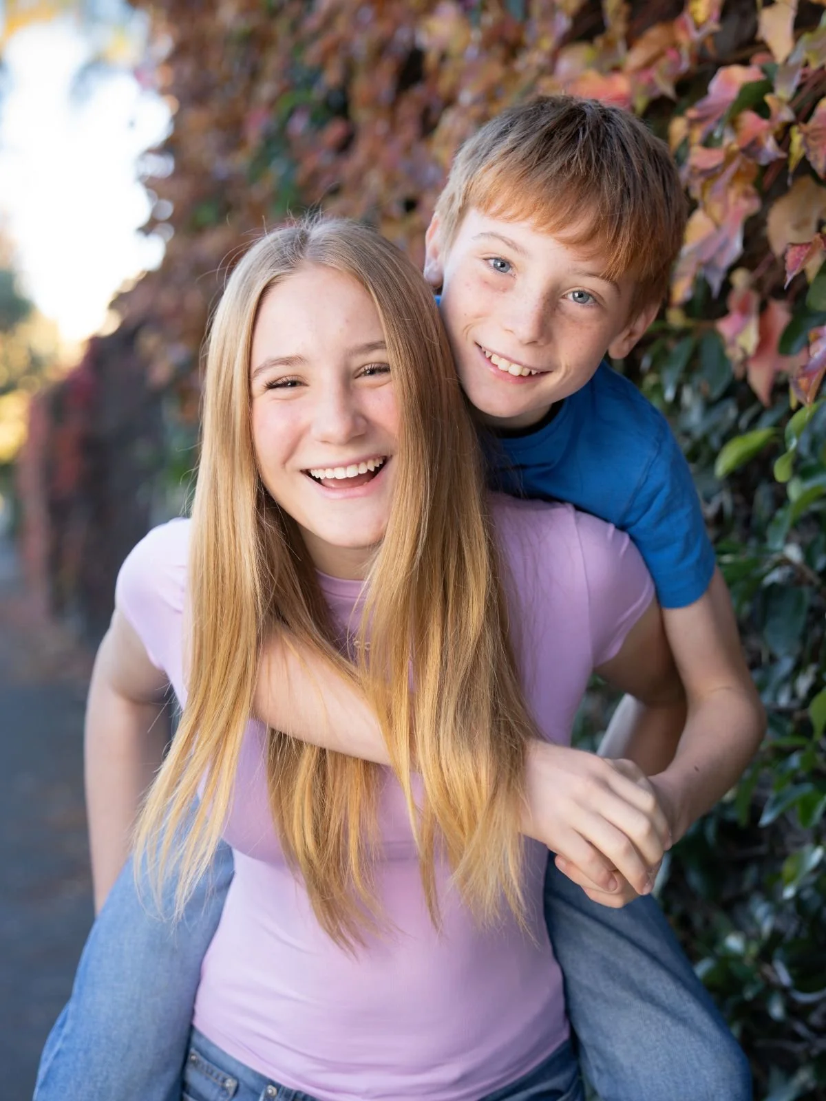 A young girl giving a piggyback ride to a boy, both smiling and outdoors, with colorful autumn leaves in the background.