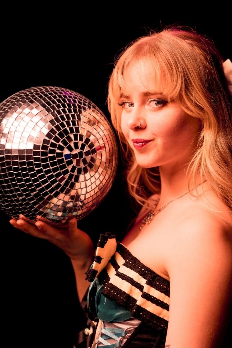 A young woman with red hair and a nose piercing holding a disco ball, posing against a black background.