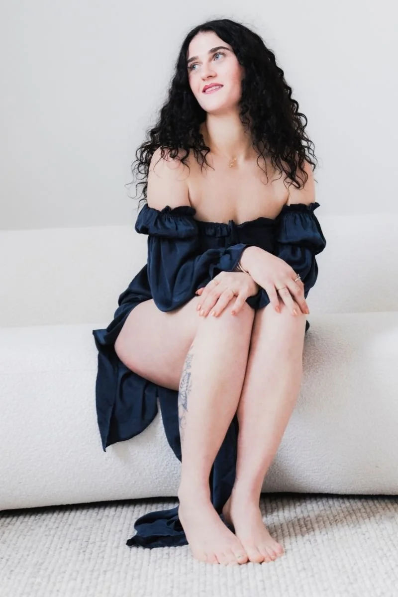 A young woman with curly dark hair and light skin sitting on a white couch with her legs crossed, wearing a navy blue off-the-shoulder dress. She is looking to the side with a slight smile, resting her hands on her knees, in a minimalistic room with beige carpet and white wall.