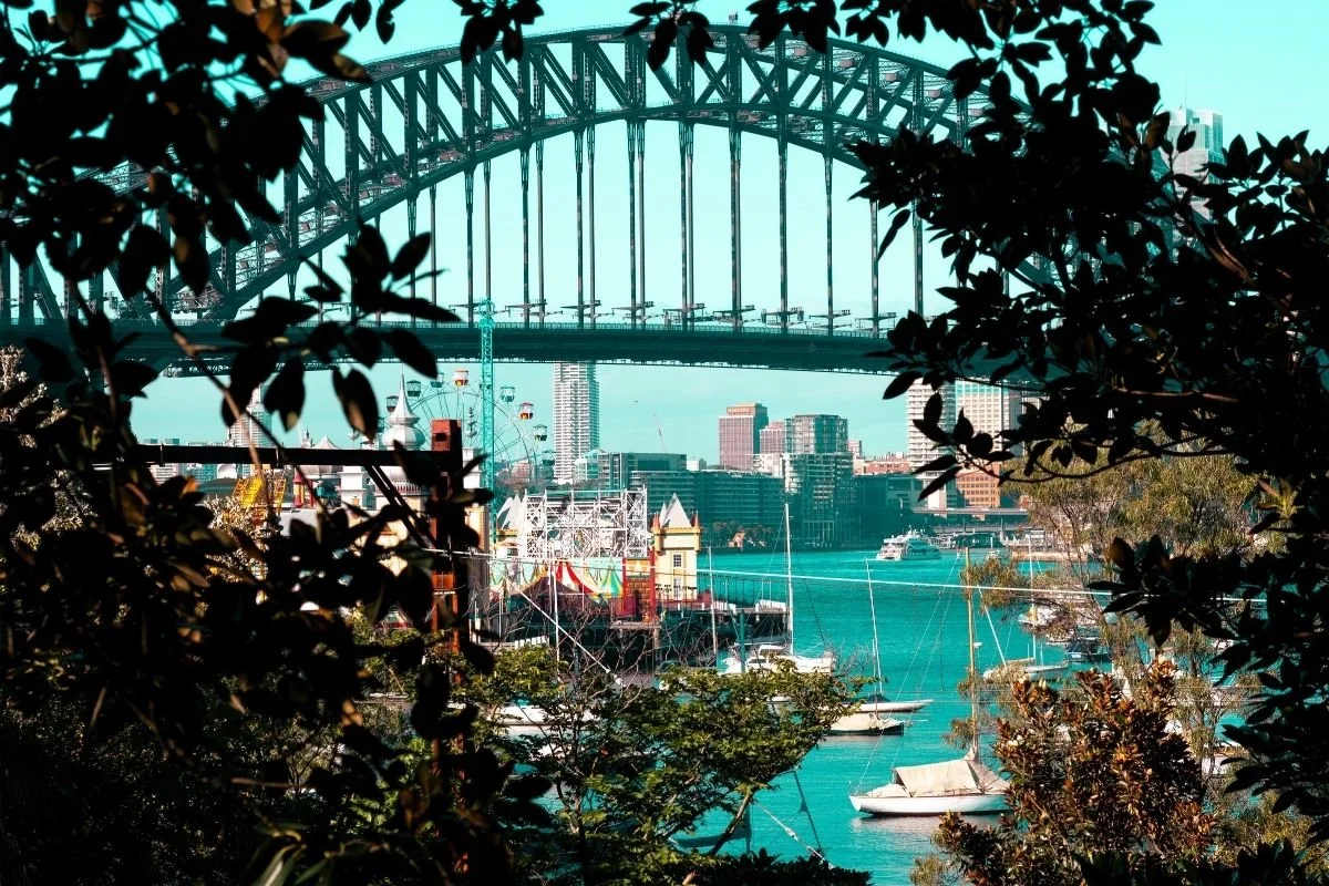 Sydney Harbour Bridge with boats on the water and a circus on the shore, framed by trees.