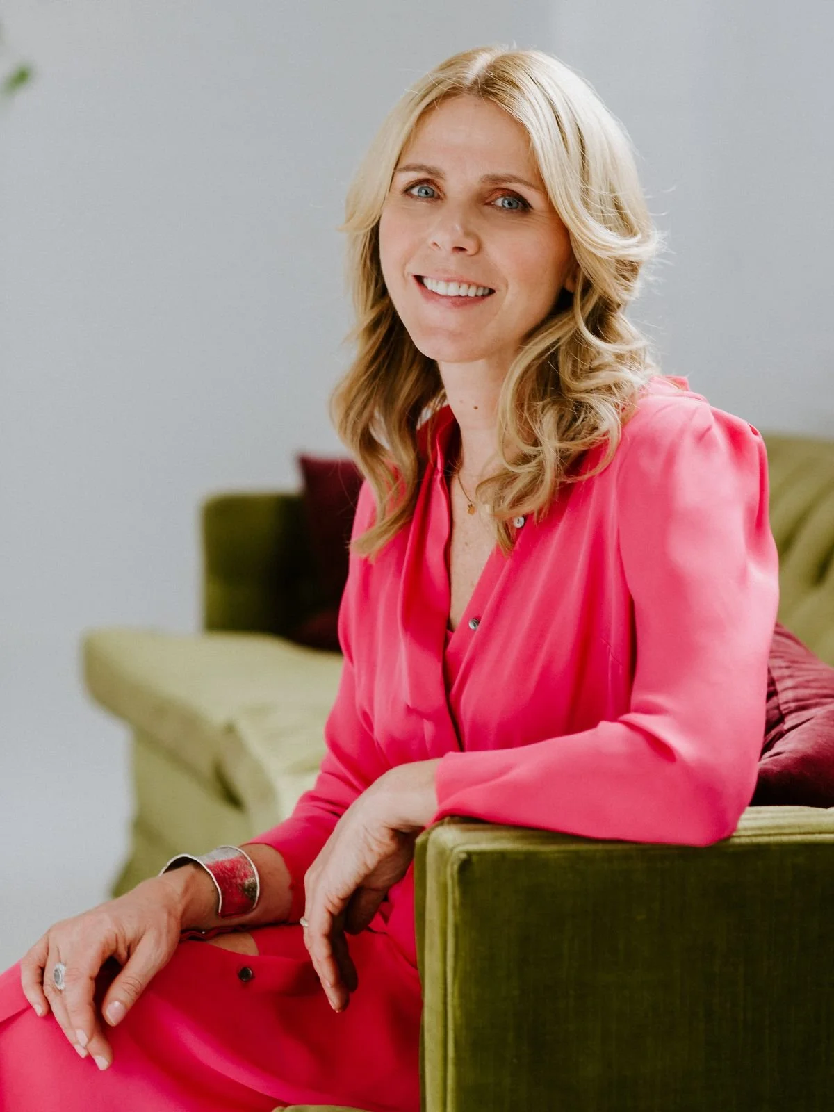 A woman with blonde hair sitting on a green couch, wearing a pink dress, smiling at the camera.