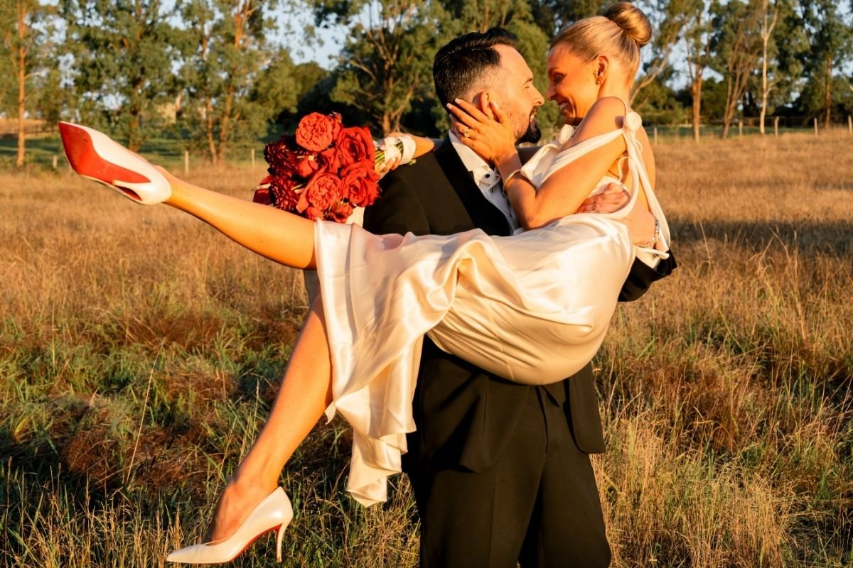 A groom in a black tuxedo holding a bride in a white wedding dress, with the bride holding a bouquet of red flowers. The bride's legs are extended, and both are smiling at each other in a field with trees in the background during sunset.
