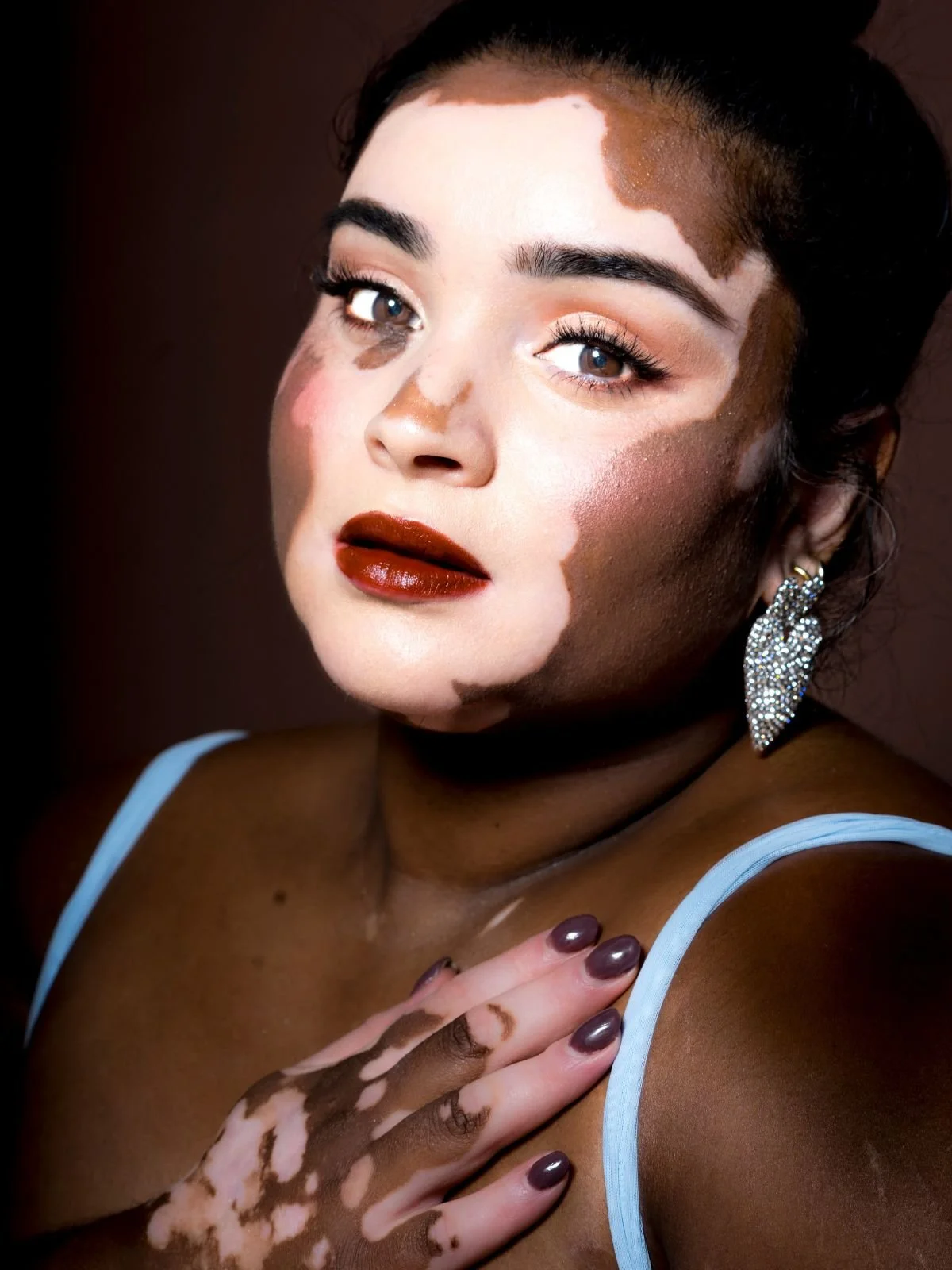 Close-up portrait of a woman with mixed skin tones, makeup, dark lipstick, and decorative eye makeup, wearing an earring and a light blue strap top, touching her shoulder with painted nails.