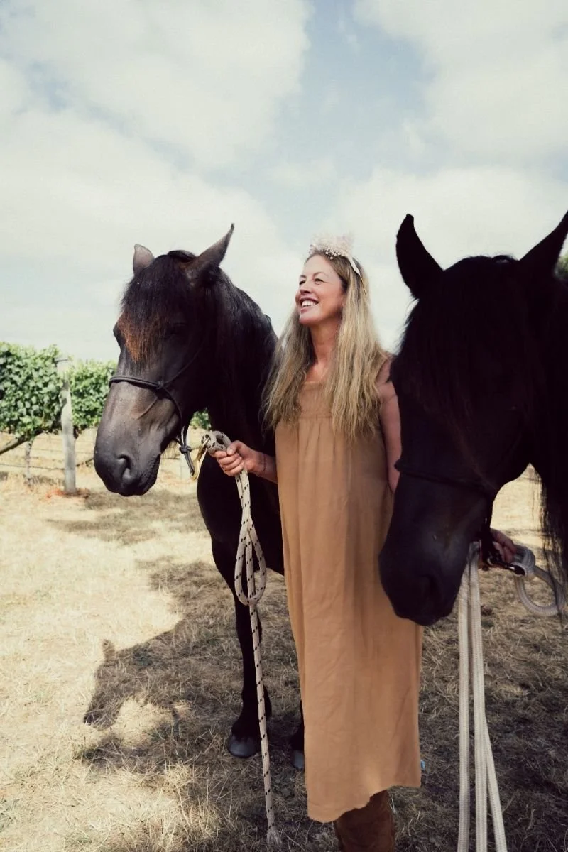 A woman with long blonde hair wearing a tan dress and a floral headband stands between two black horses outdoors, smiling at each other. The scene is set under a partly cloudy sky with a vineyard in the background.