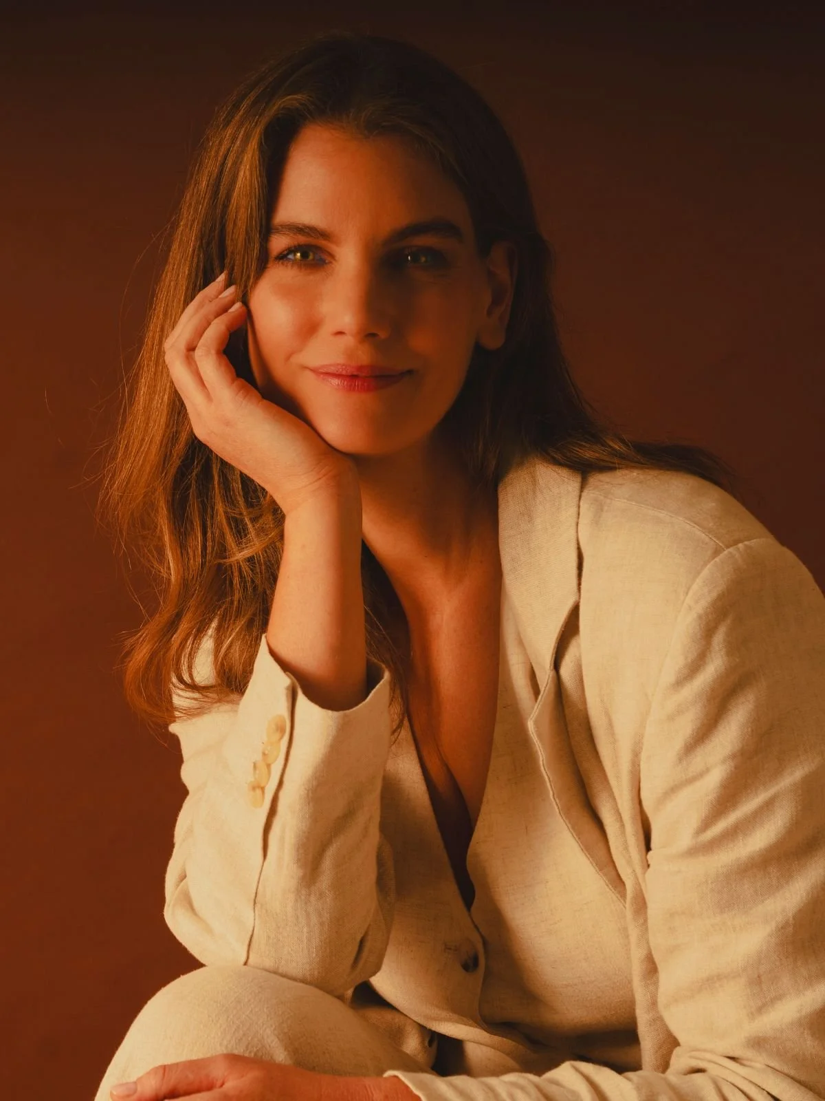 A woman with shoulder-length brown hair, wearing a beige blazer, sitting and resting her head on her hand, smiling softly at the camera against a warm, brown background.