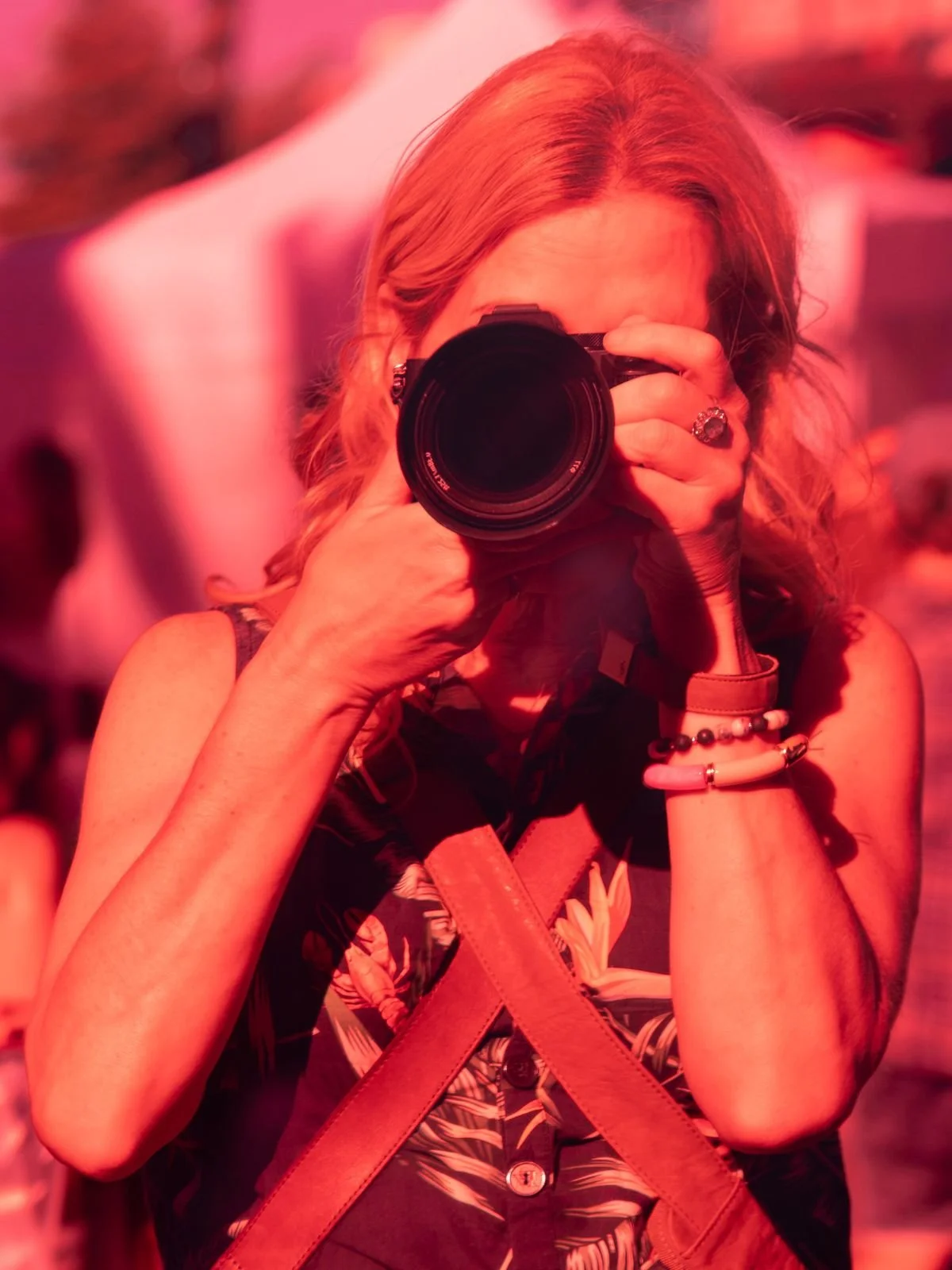 Woman with long hair taking a photograph with a camera, wearing bracelets and a floral shirt, in a setting illuminated with pinkish-red light.