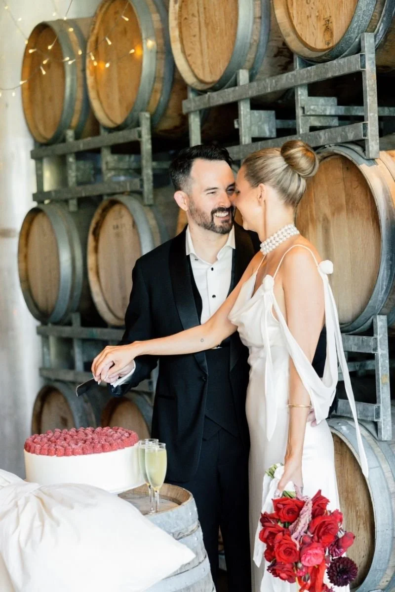 A newlywed couple sharing a romantic moment behind a wedding cake at a wine cellar, with wine barrels stacked in the background.