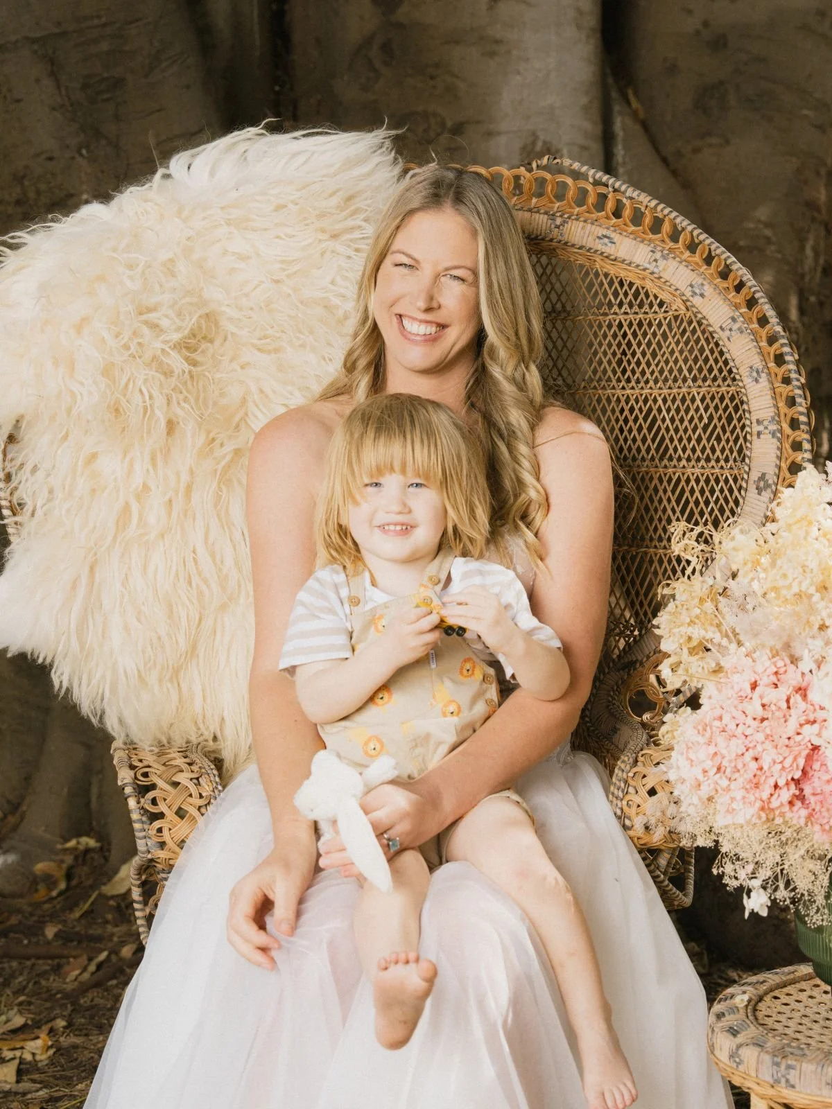 A woman and a young child sitting together on a rattan chair in an outdoor setting. The woman has long, wavy blonde hair and is wearing a white dress, smiling and winking. The child, with red hair and wearing a beige outfit with a pattern, has a playful expression and is holding a small toy. There are flowers and a sheepskin throw beside them.