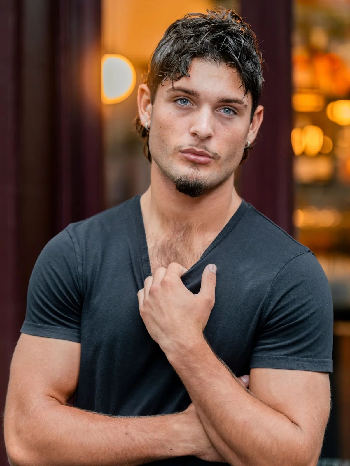 A young man with blue eyes, dark hair, and earrings, wearing a dark gray t-shirt, posing outdoors with his hand near his chest and head slightly tilted.