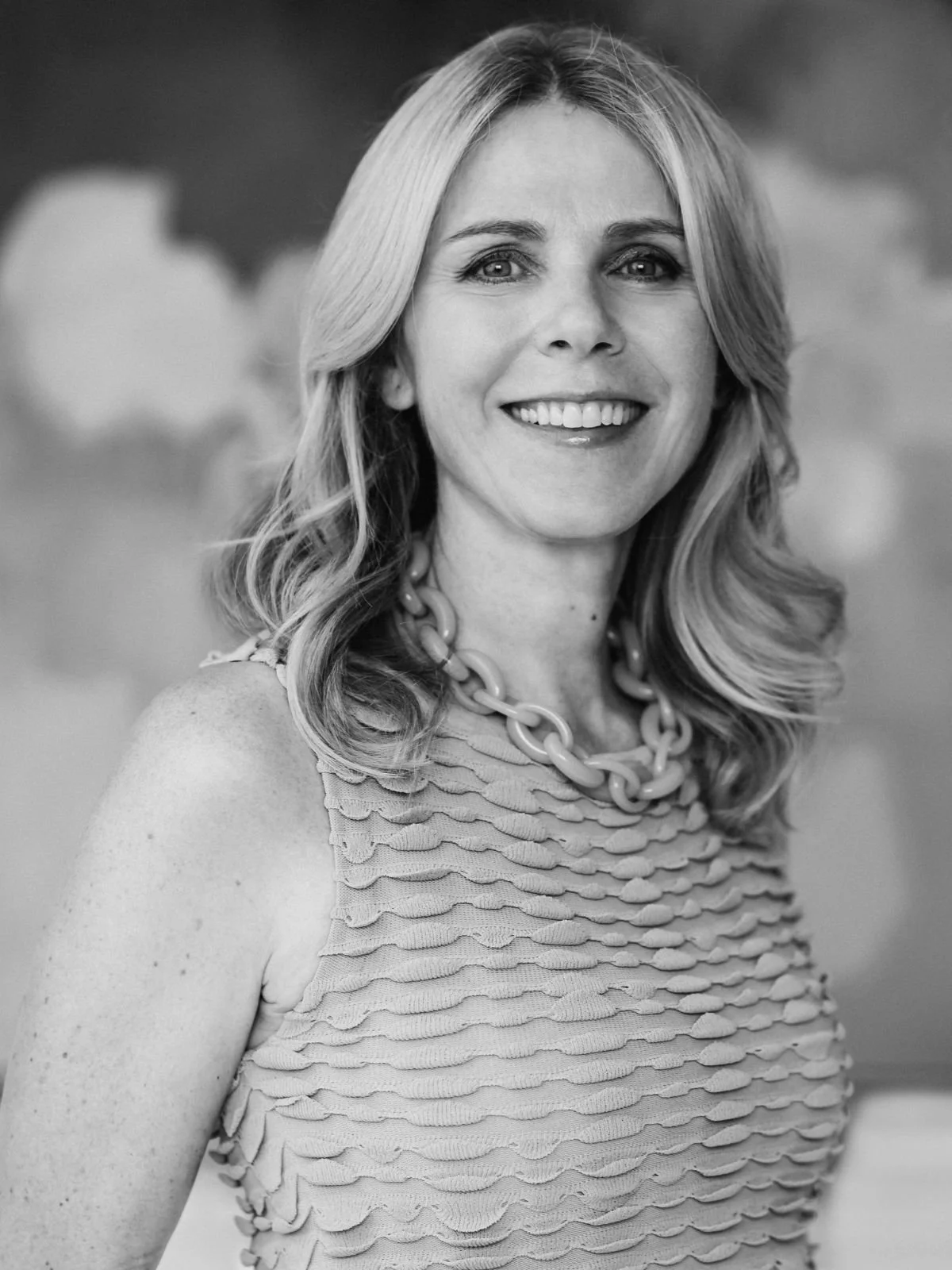 Black and white photo of a woman with wavy, shoulder-length hair, wearing a textured sleeveless top and a chunky necklace, smiling at the camera.