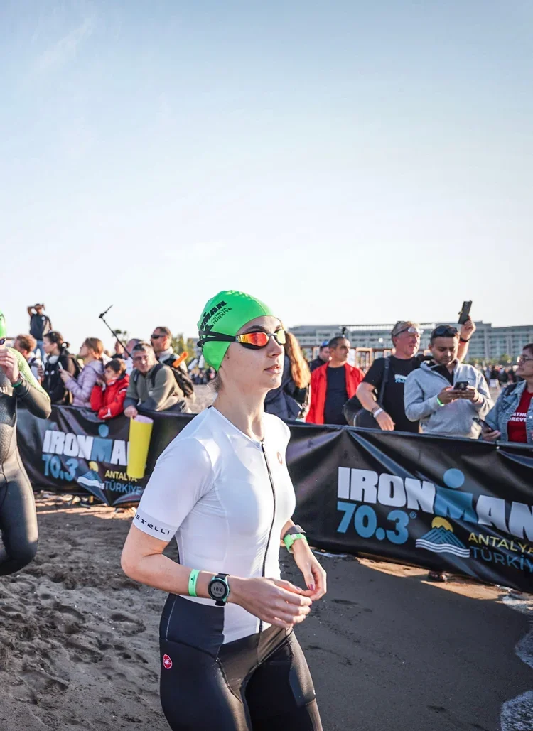 Female athlete wearing a green swim cap, sunglasses, and a white and black wetsuit at the Ironman 70.3 race event with spectators and banners in the background.