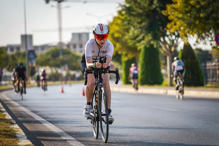 A cyclist in a white jersey and helmet riding on a paved street with a few other cyclists in the background, some orange traffic cones, and trees along the sidewalk.