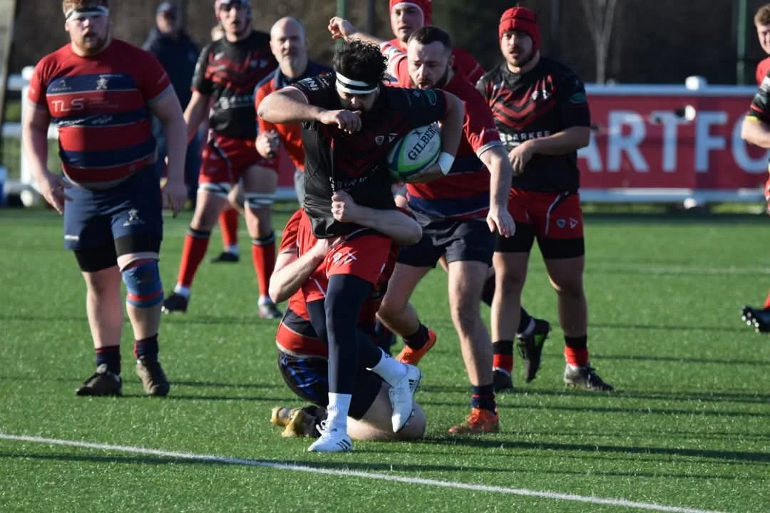 Rugby players in action with one player catching the ball while being tackled by opponents on a grass field.