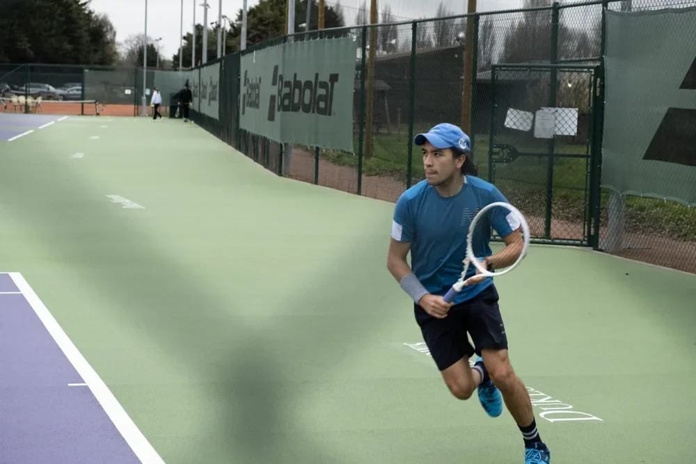 Tennis player in blue shirt and cap running on tennis court with racket in hand.