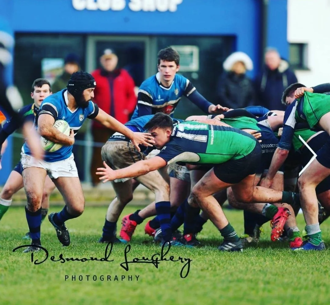 A rugby game with players in blue and green jerseys engaged in a scrum, some players are pushing and others are holding the ball, with a blue club shop building in the background.
