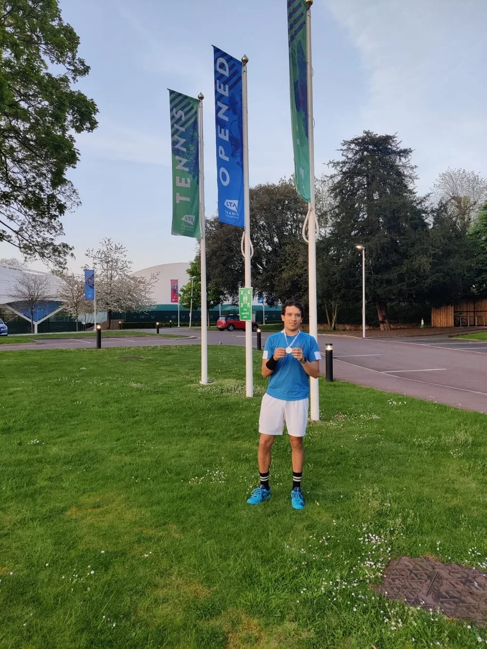 A young man standing on a grassy area holding a medal in front of three flagpoles at a tennis venue, with trees and a parking lot in the background.