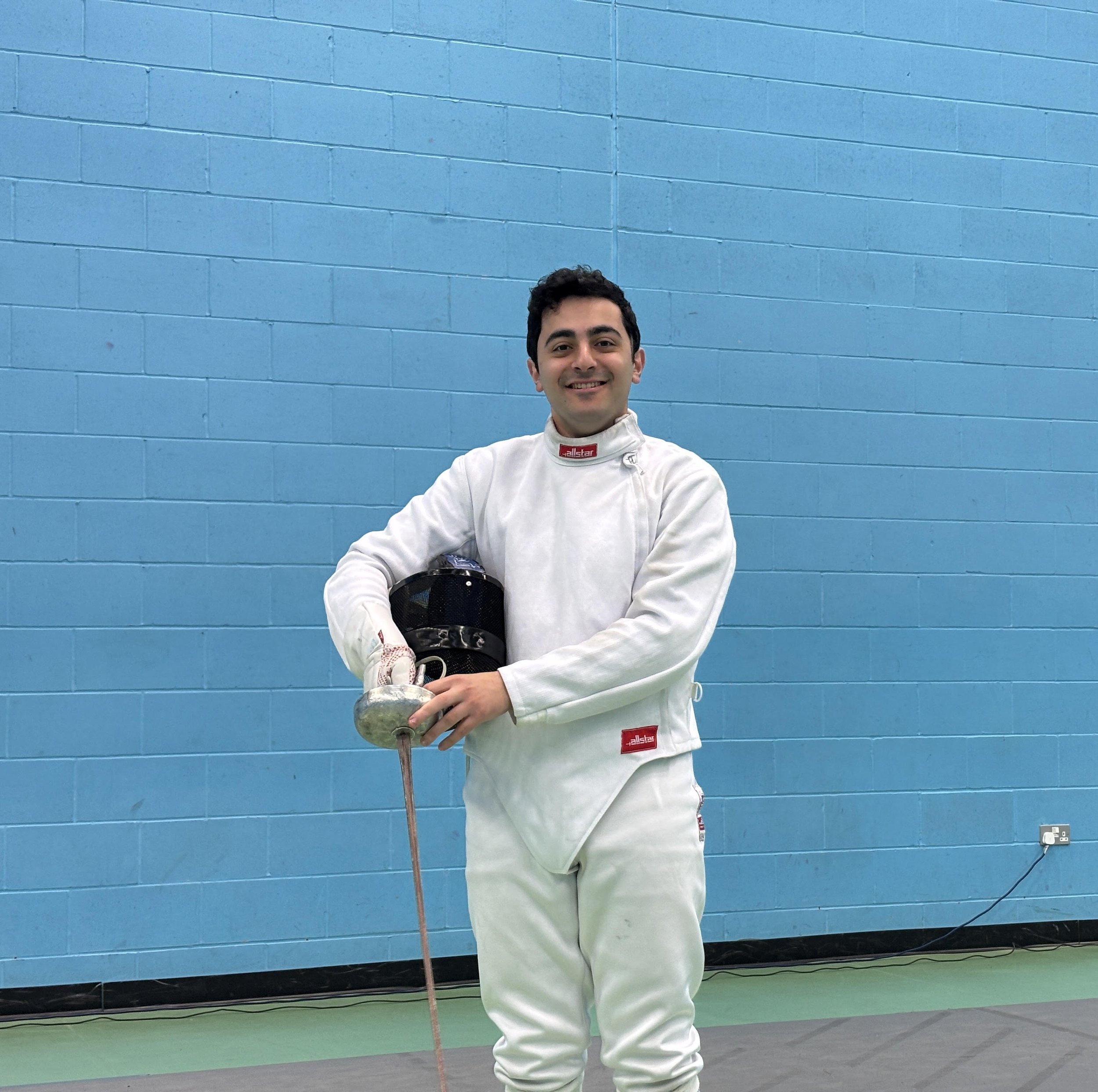 A smiling man in fencing attire, holding a fencing mask and a foil, standing against a blue brick wall in a gym or sports hall.