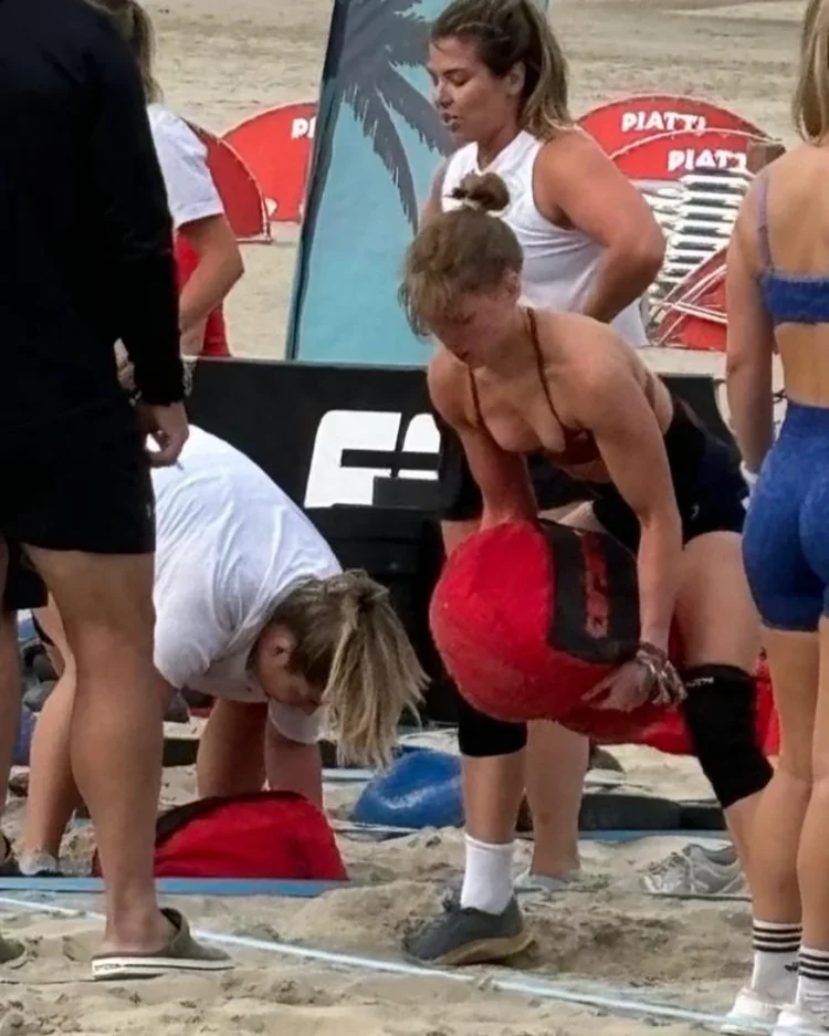 Women and children on a sandy beach, some engaging in fitness activities with exercise bags and equipment, near U.S. Coast Guard banners.
