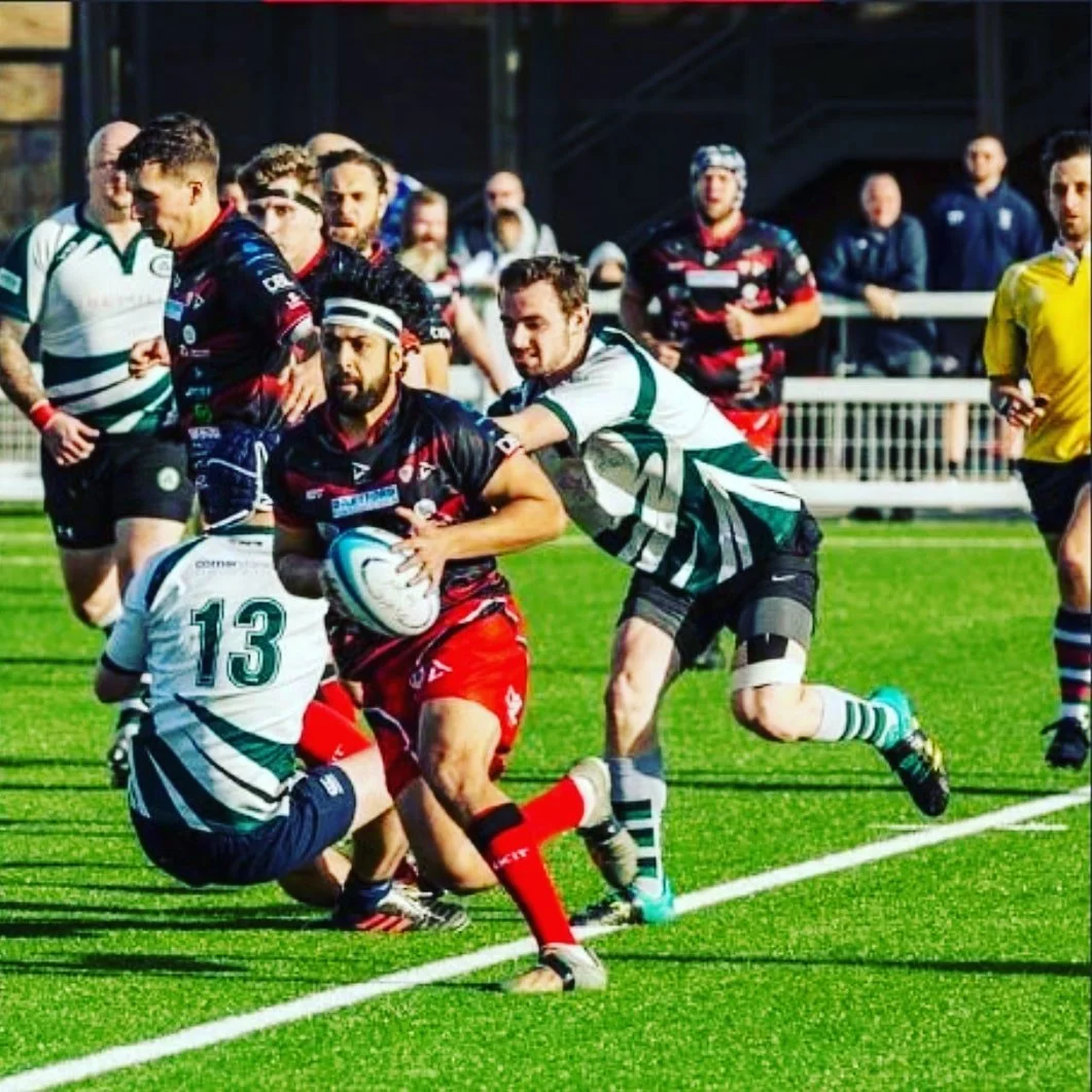 Rugby players on the field during a match, with one player holding the ball while being tackled by an opponent, and other players in the background.