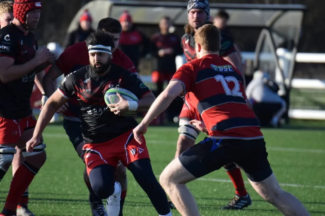 A rugby game with players in red and black uniforms. One player is holding the ball and running, while another player is preparing to tackle.