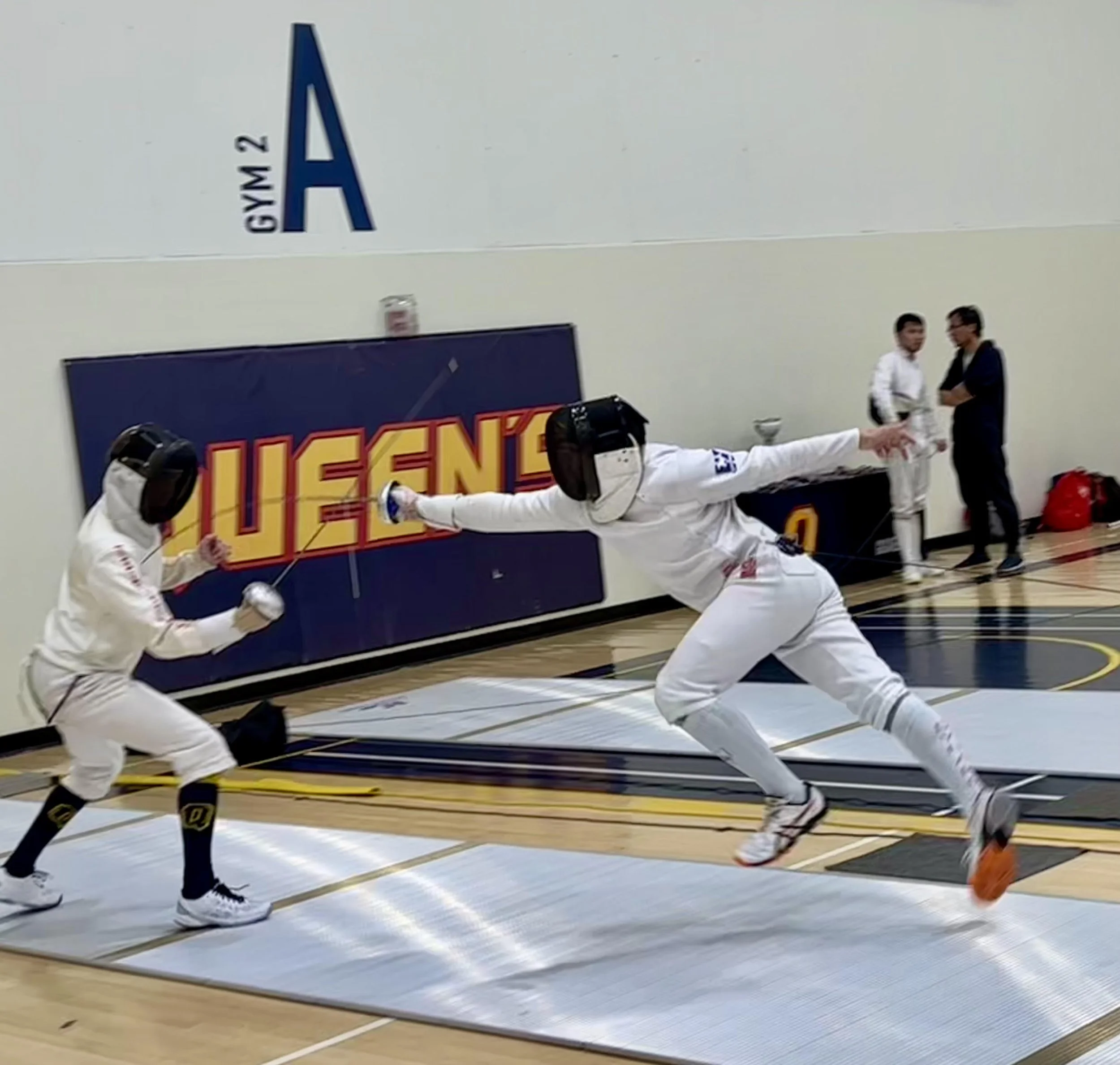 Two fencers engaged in a duel, wearing white protective gear and masks, on a fencing piste with an electronic scoring system, with two men watching in the background.