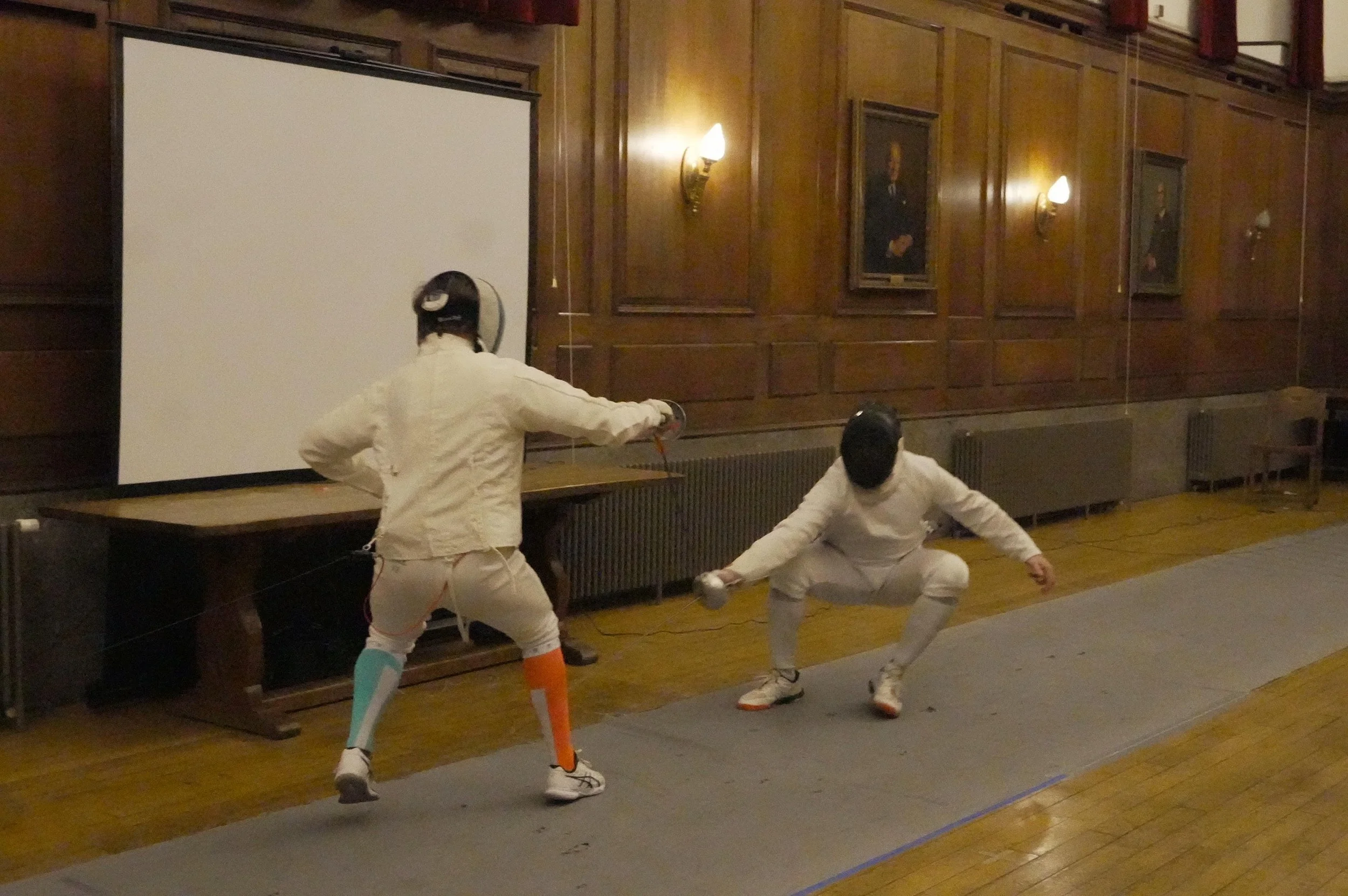 Two fencers in white uniforms and protective gear engaging in a fencing match indoors. One is lunging forward with an extended arm, while the other is in a defensive position. The room has wooden walls with framed portraits, wall lamps, and a large w