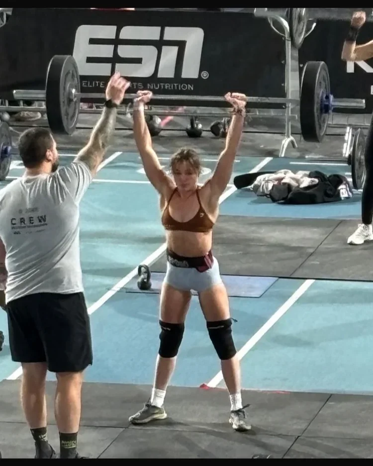 A woman lifting a barbell overhead during a weightlifting competition at a gym. She is wearing a sports bra, shorts, knee sleeves, and athletic shoes. A man in a t-shirt and shorts is spotting her, standing nearby.