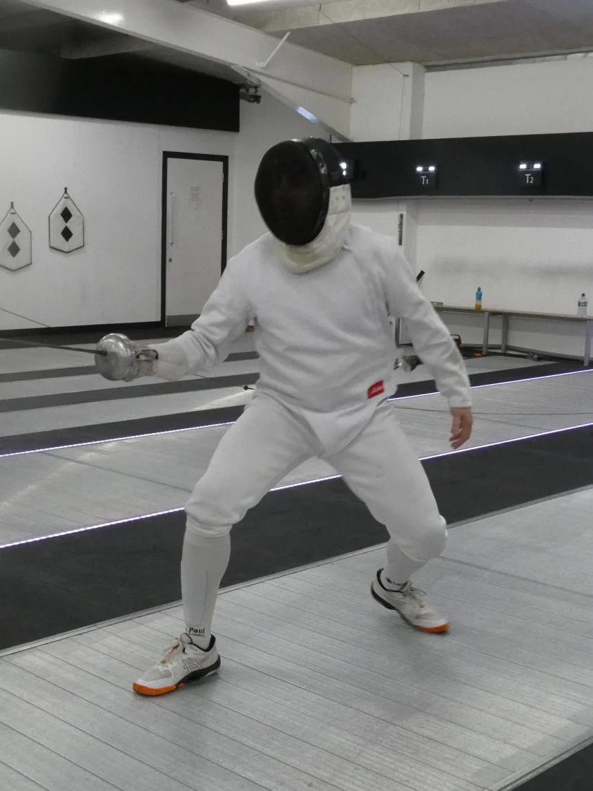 Person dressed in white fencing gear practicing fencing in a modern indoor fencing facility.