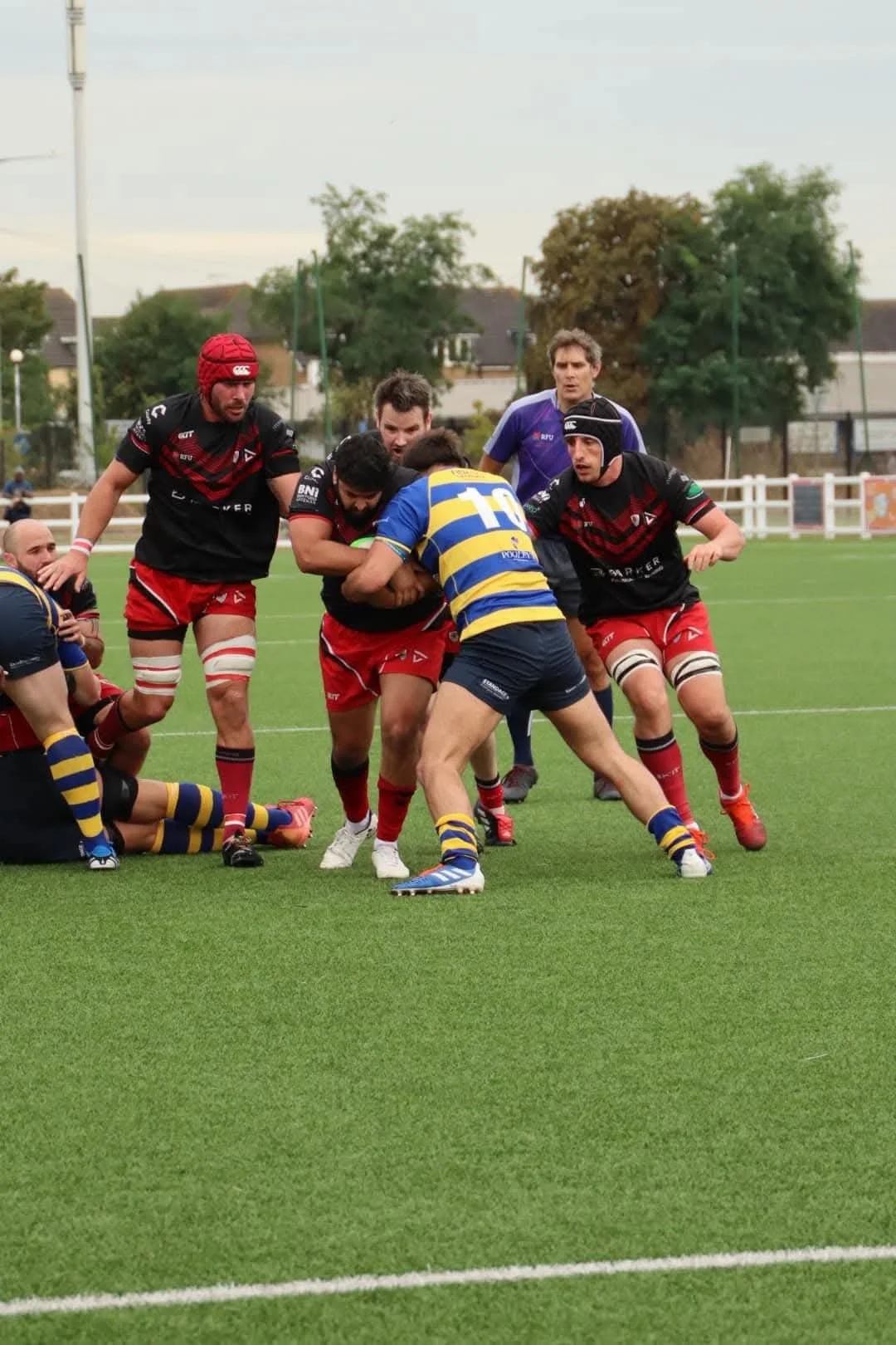 Rugby players engaged in a tackle during a match on a grassy field with trees and houses in the background.
