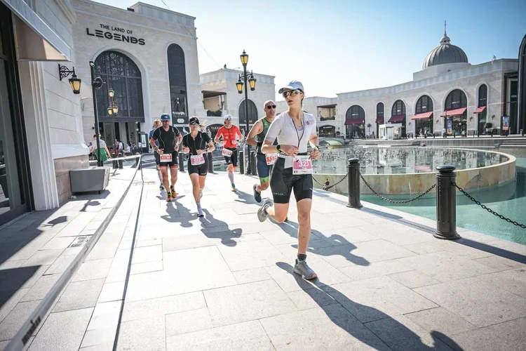 Group of runners participating in a race along a city street with water feature, hotels, and shops in the background.