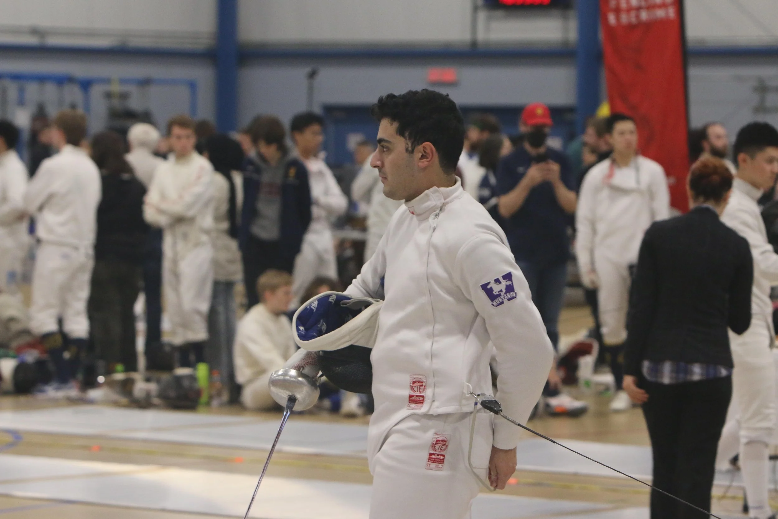 A young male fencer in a white fencing uniform holding a fencing sword, standing among other competitors and spectators in an indoor sports arena.
