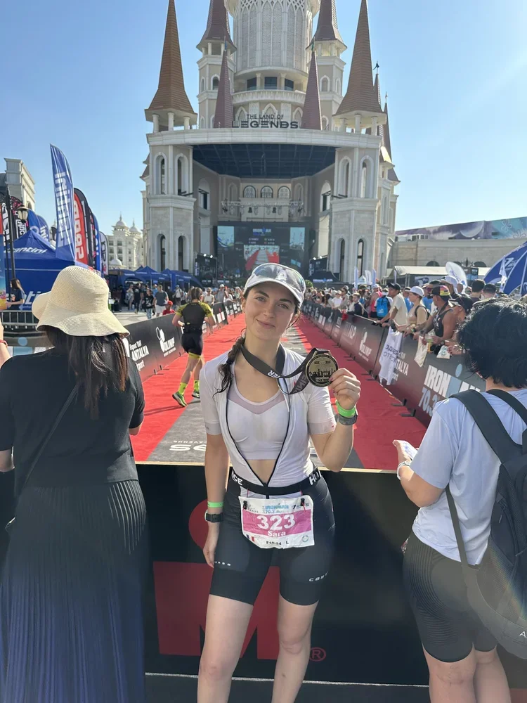 A young woman holding a marathon medal at the finish line of an Ironman race in front of a castle-themed building at Disneyland, with a crowd celebrating around her.