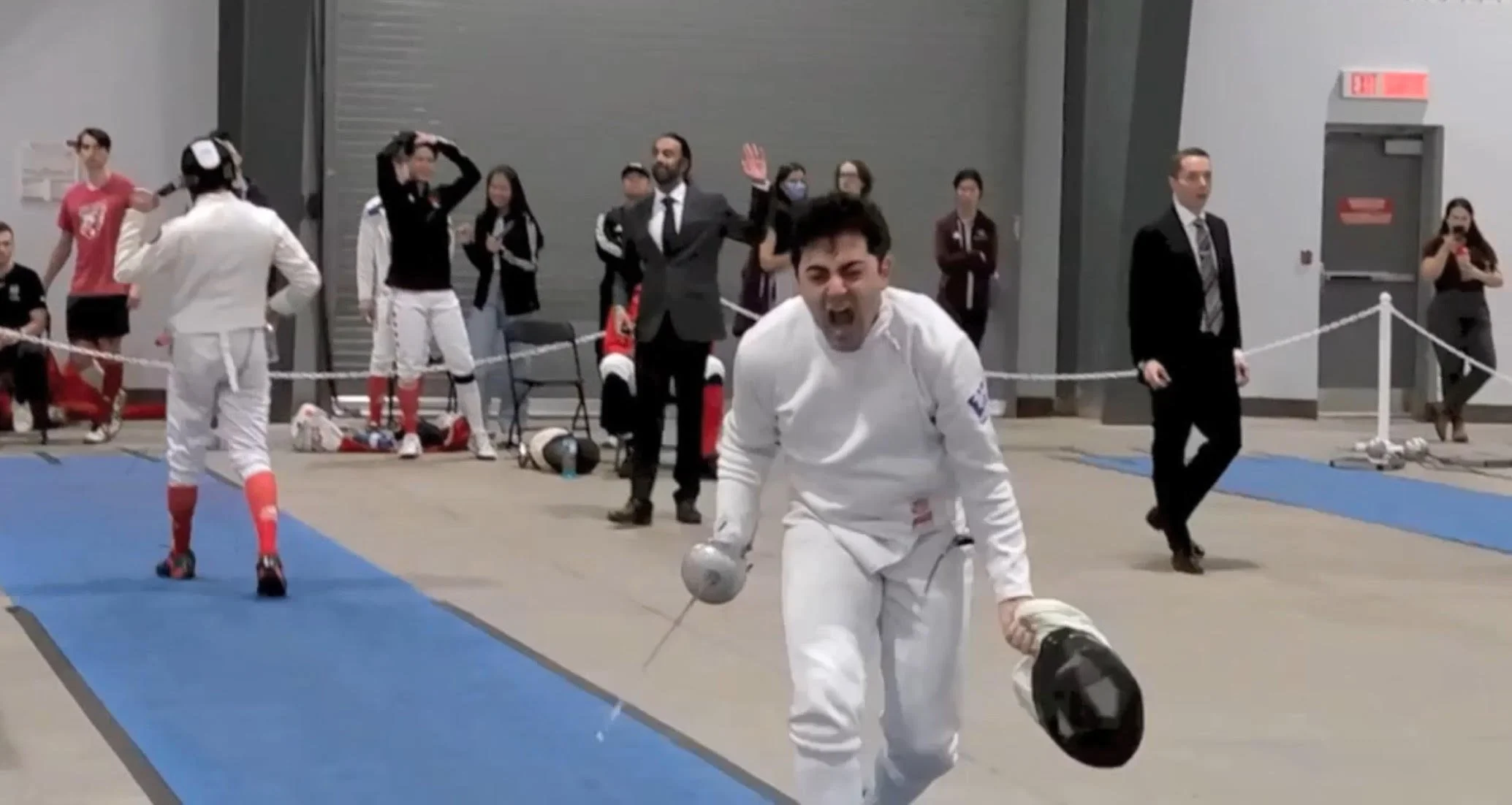 Fencer in white uniform jumping and falling on a blue fencing strip during a fencing competition, with spectators and officials watching in the background.