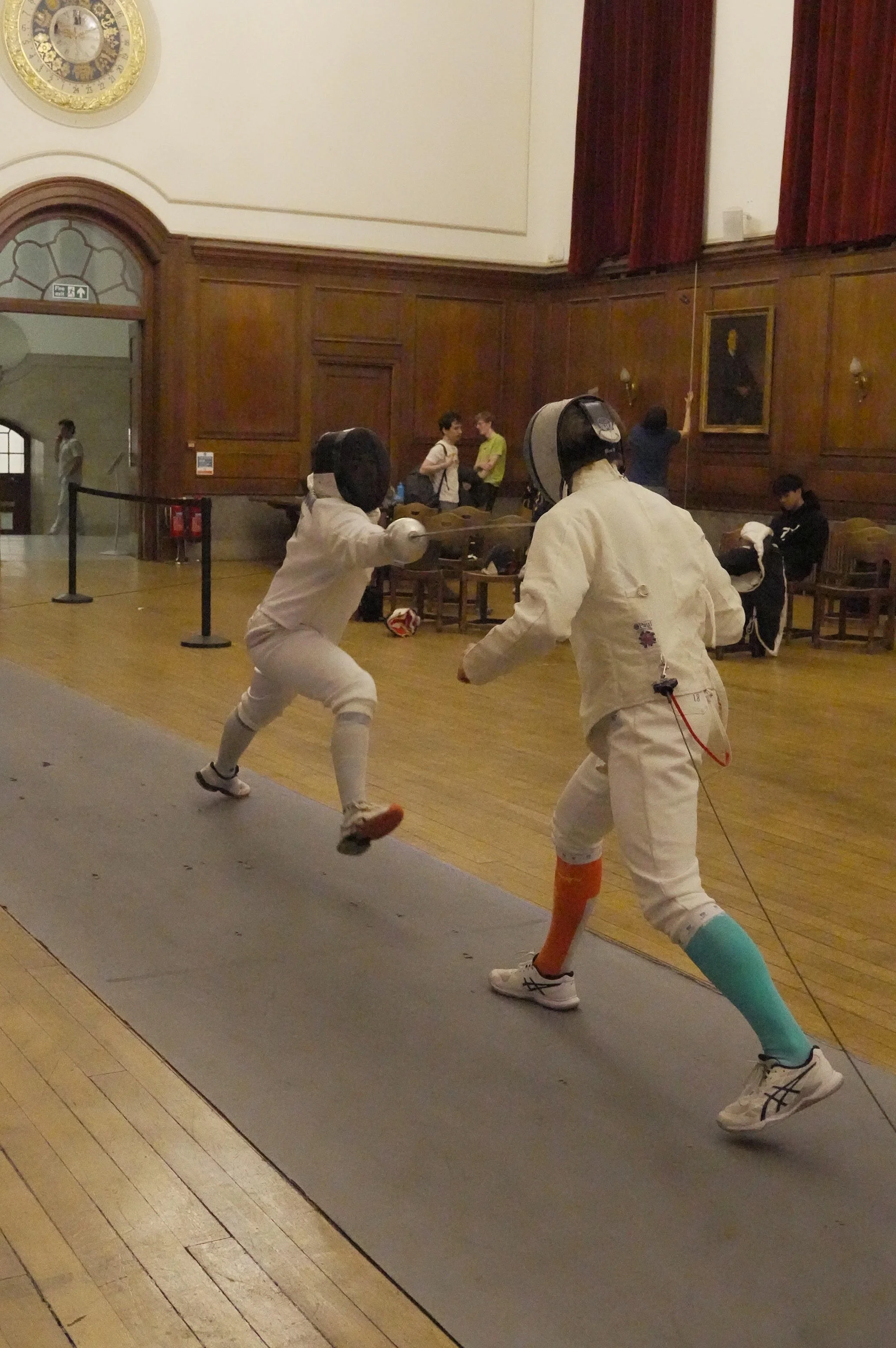 Two fencers in white uniforms and protective masks are engaged in a fencing match on a gray fencing strip inside a room with wooden walls and red curtains. Several people are watching in the background.