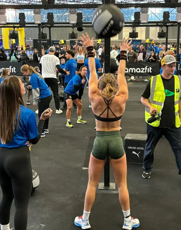 Woman with a muscular back wearing a black sports bra and green shorts lifting a heavy medicine ball in a gym during a fitness event.