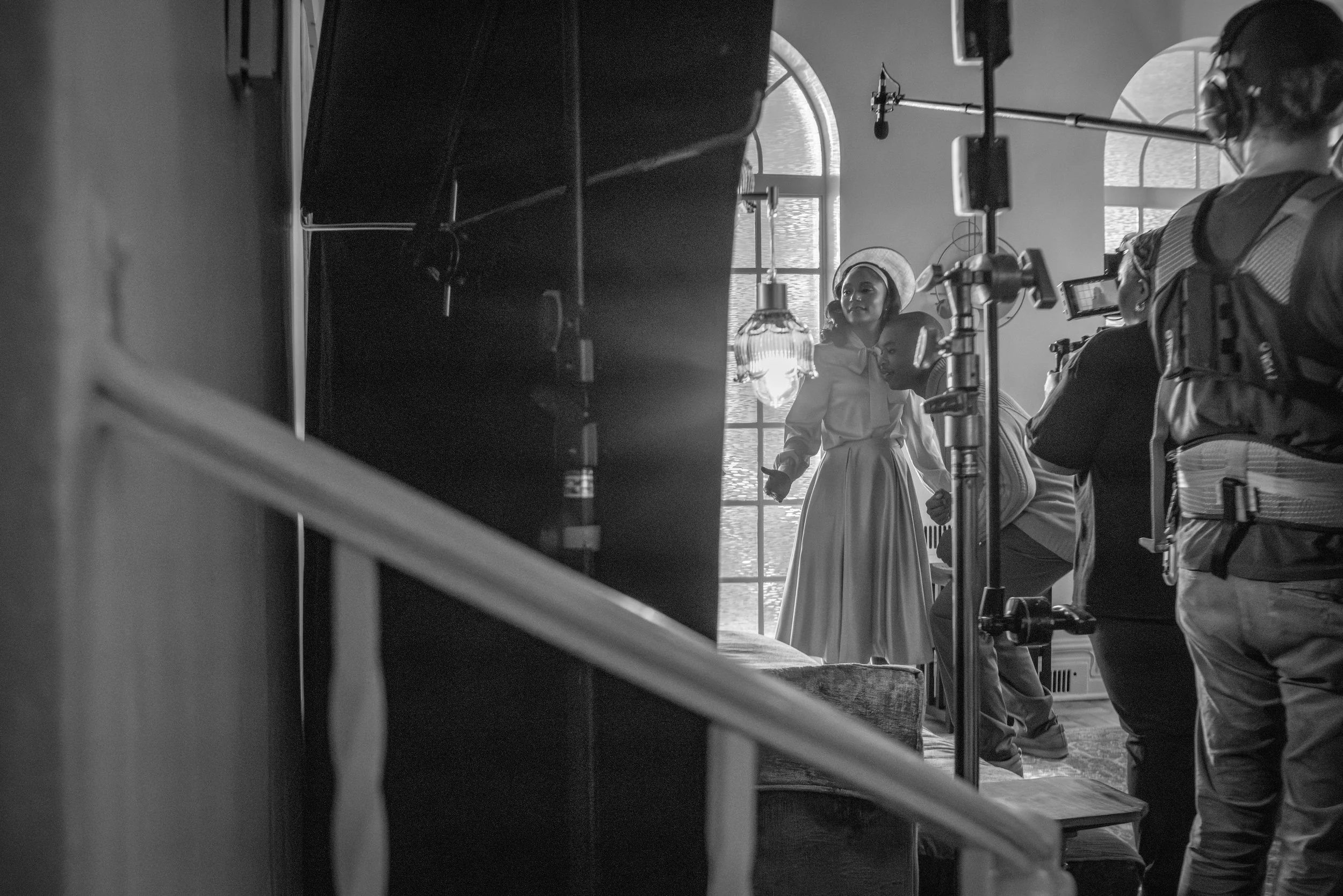 Black and white photo of a woman in vintage attire with a hat, standing in a room filled with crew members, equipment, and lights, during a film or television shoot.