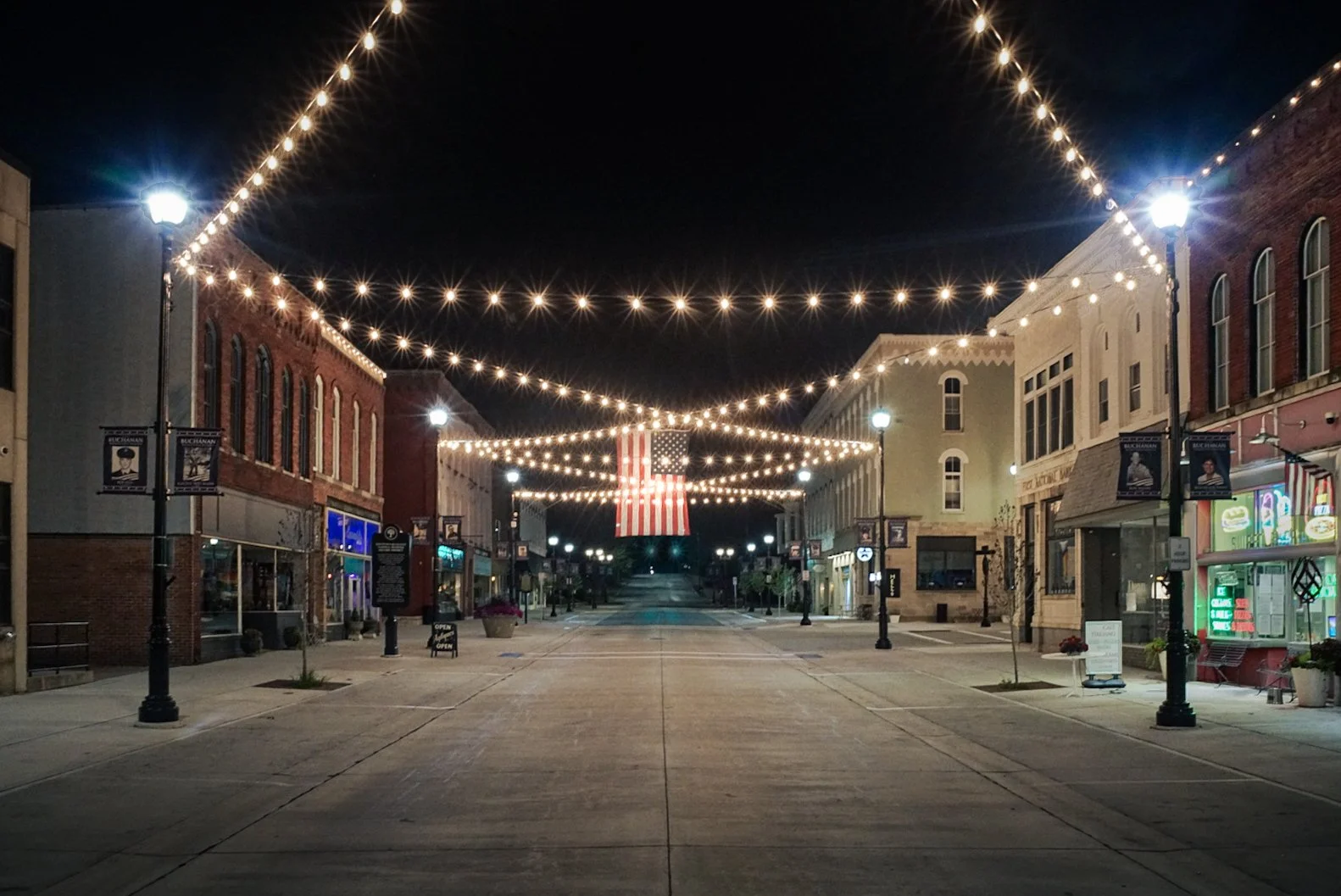 Empty downtown street at night decorated with string lights and an American flag hanging in the center.