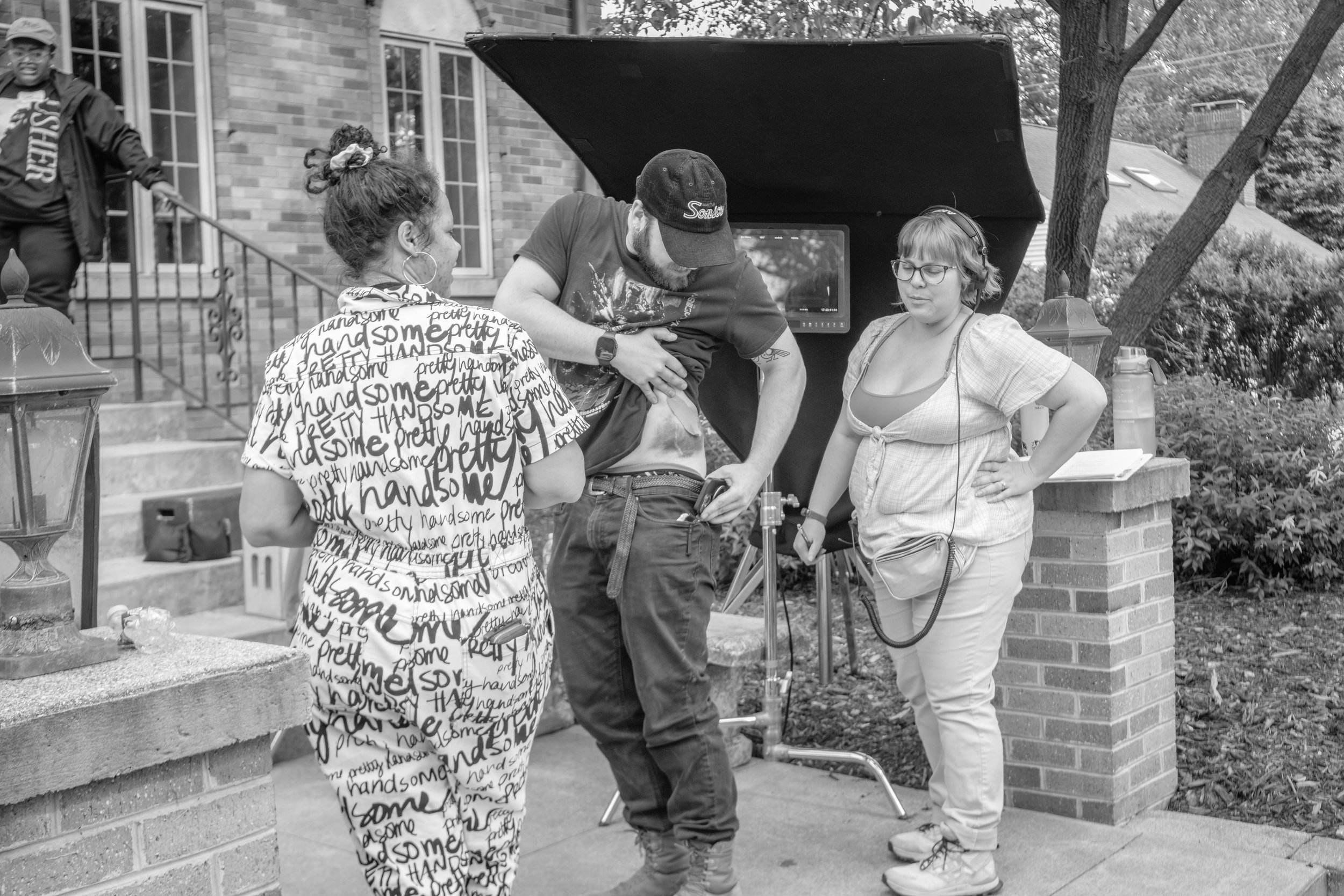 A film crew on a residential front porch with brick columns, surrounded by trees. Three crew members are visible, including a woman with headphones and glasses holding a monitor, a man with a cap and beard focusing on a camera, and a woman in a shirt