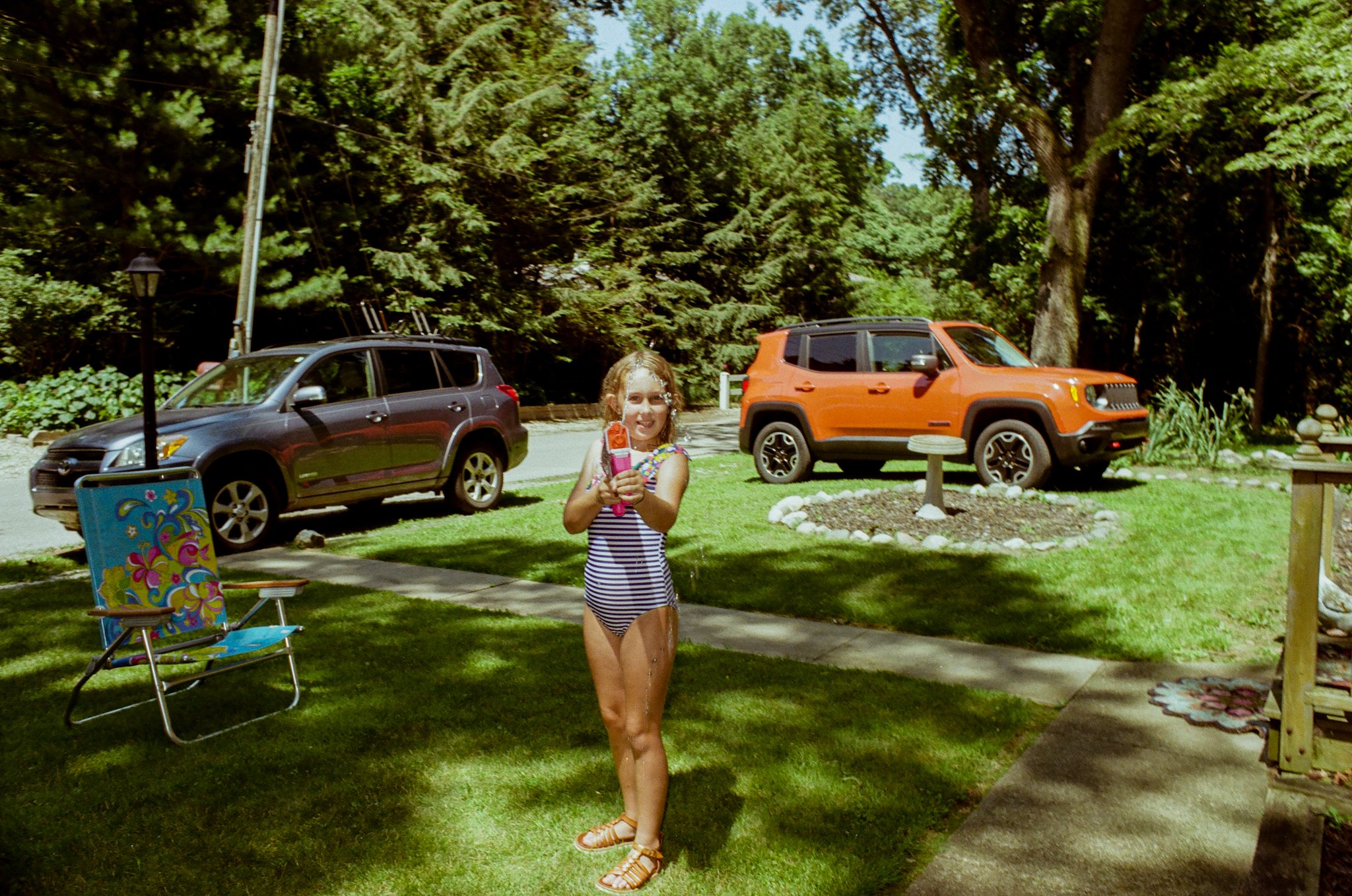 A young girl in a striped swimsuit is smiling and holding a water gun. She is outdoors in a grassy yard with two parked cars, trees, and a small paved sidewalk nearby.