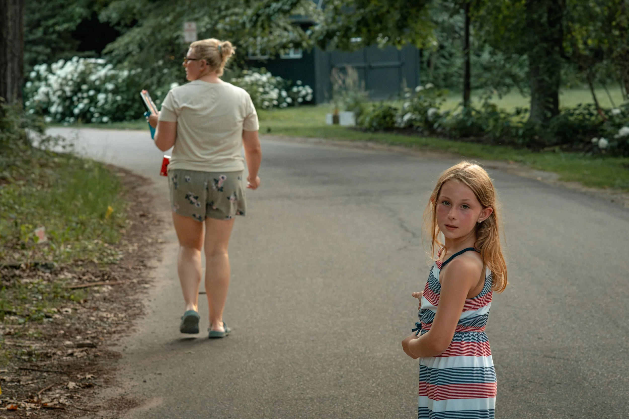Two young girls walking on a residential street surrounded by greenery, with one girl looking at the camera and the other girl walking ahead holding a book.